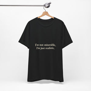 The "I'm Not Miserable. I'm Just Realistic." T-Shirt by Printify (Old Man Snark) is a black crew neck made from soft Airlume cotton, shown hanging on a wooden hanger against a white background with beige text.