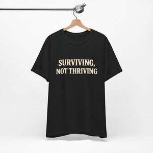 A "Surviving. Not Thriving." T-Shirt by Old Man Snark from Printify, made of soft Airlume cotton, hangs on a wooden hanger, displaying bold beige lettering—a nod to modern life.