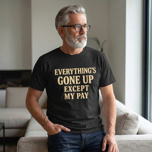 A gray-haired, bearded man in glasses stands by a sofa in a bright living room, wearing the Printify "Everything's Gone Up Except My Pay" T-Shirt by Old Man Snark, reflecting on rising living costs as he looks out the window.
