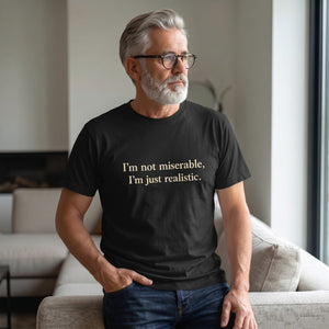 A gray-haired, bearded man in glasses wears the Printify "I'm Not Miserable. I'm Just Realistic." T-Shirt by Old Man Snark as he sits on a sofa by a large window, gazing thoughtfully into the distance.
