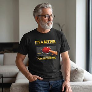 A gray-haired, bearded man in glasses wears the Printify "It's A Button. Push The Button." T-Shirt by Old Man Snark, featuring a red button graphic, while standing in a modern living room.