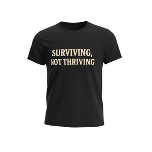The "Surviving. Not Thriving." T-Shirt by Printify (Old Man Snark) features bold beige letters on soft Airlume cotton, showcased on a plain white background.