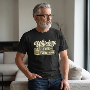 A gray-haired, bearded man in glasses stands by a window, wearing the "Whiskey Fixes Everything" T-Shirt by Printify—an ideal men's gift—resting his hand on a sofa and gazing thoughtfully outside.