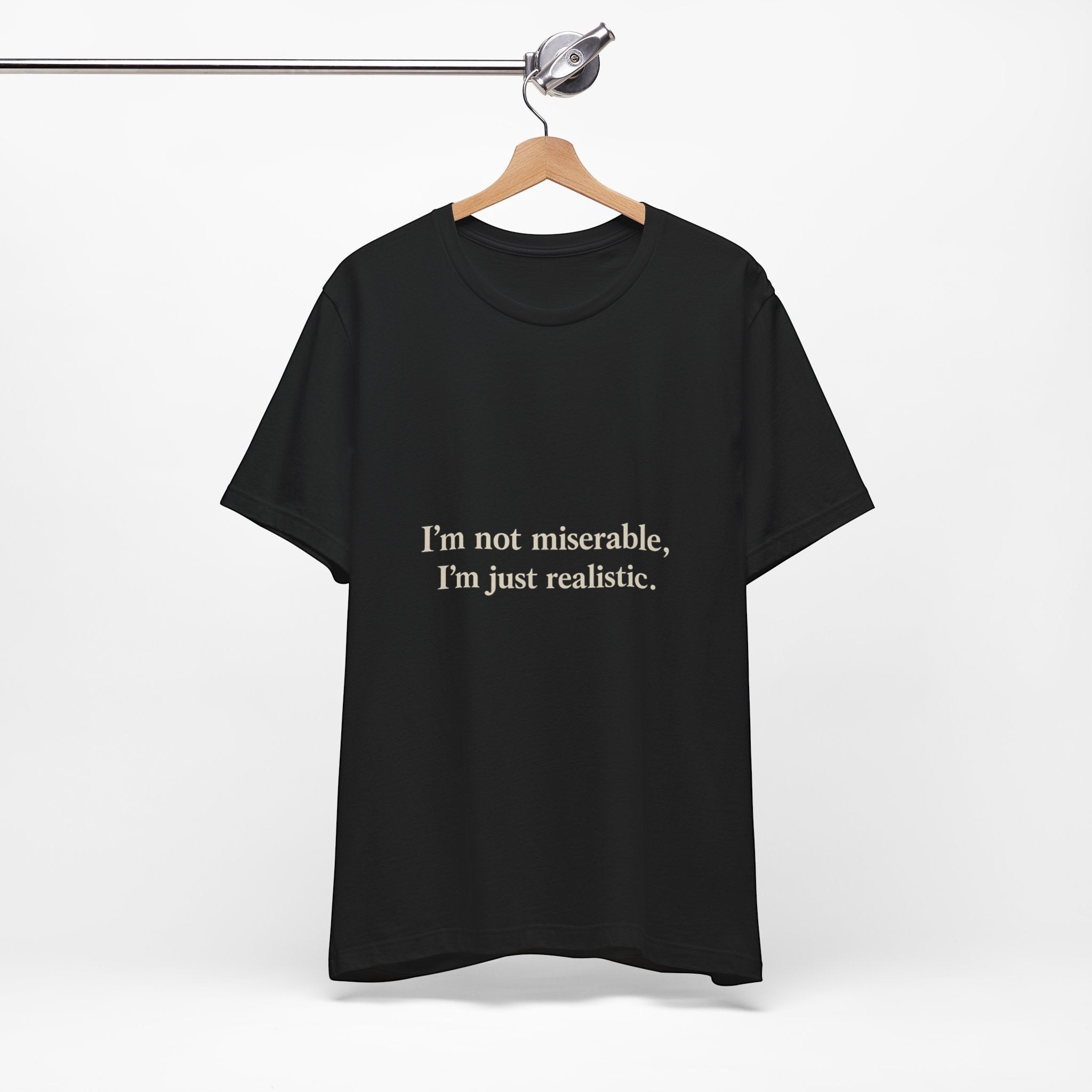 The "I'm Not Miserable. I'm Just Realistic." T-Shirt by Printify (Old Man Snark) is a black crew neck made from soft Airlume cotton, shown hanging on a wooden hanger against a white background with beige text.