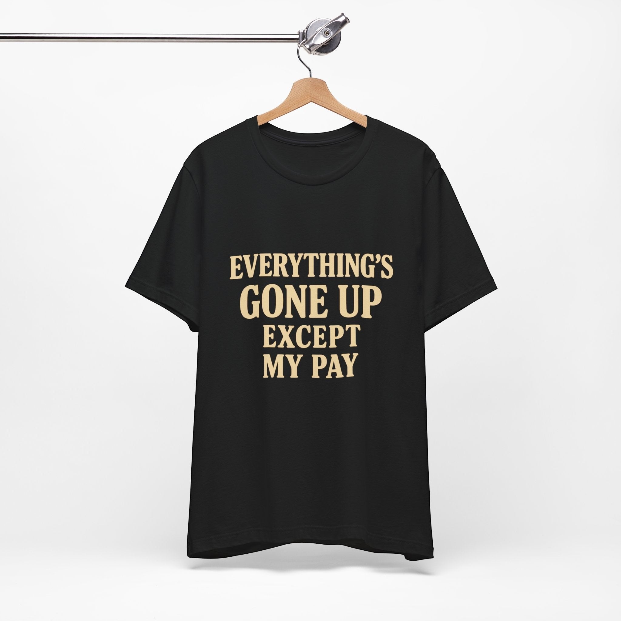 The "Everything's Gone Up Except My Pay" T-Shirt by Printify (Old Man Snark) features bold beige lettering on black Airlume cotton, displayed on a wooden hanger against a white background.