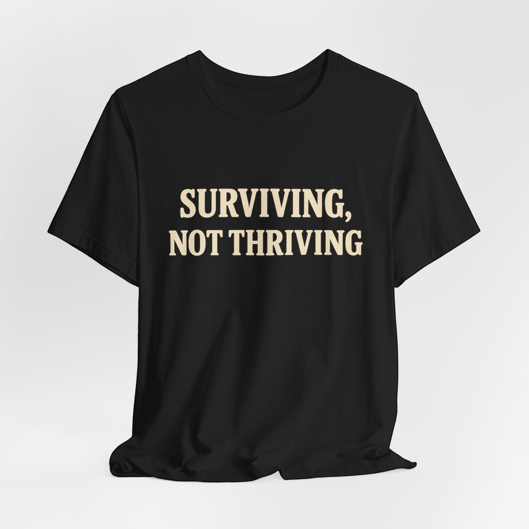 The "Surviving. Not Thriving." T-shirt by Old Man Snark from Printify is a black tee with bold beige text, crafted from soft Airlume cotton and displayed on a plain white background.