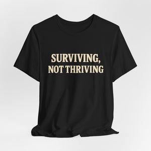 The "Surviving. Not Thriving." T-shirt by Old Man Snark from Printify is a black tee with bold beige text, crafted from soft Airlume cotton and displayed on a plain white background.