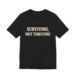 The "Surviving. Not Thriving." T-shirt by Printify is a black tee from Old Man Snark, crafted with soft Airlume cotton and bold beige lettering—a modern take on life's challenges.