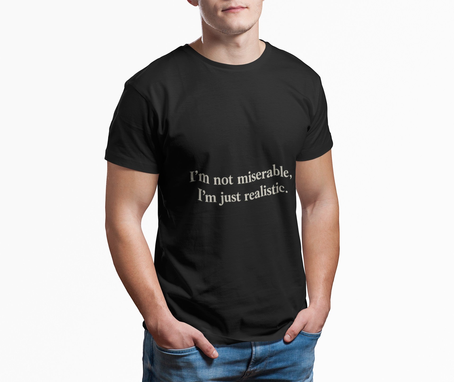A man stands with his hands in his jeans pockets, wearing the Printify "I'm Not Miserable. I'm Just Realistic." T-Shirt by Old Man Snark—black Airlume cotton with the phrase in white—against a plain white background.