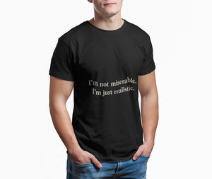 A man stands with his hands in his jeans pockets, wearing the Printify "I'm Not Miserable. I'm Just Realistic." T-Shirt by Old Man Snark—black Airlume cotton with the phrase in white—against a plain white background.