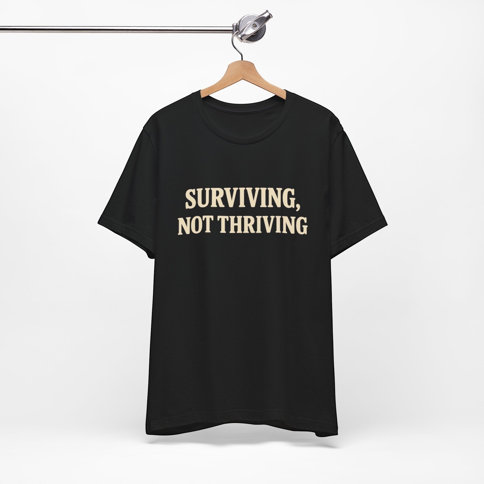 A "Surviving. Not Thriving." T-Shirt by Old Man Snark from Printify, made of soft Airlume cotton, hangs on a wooden hanger, displaying bold beige lettering—a nod to modern life.