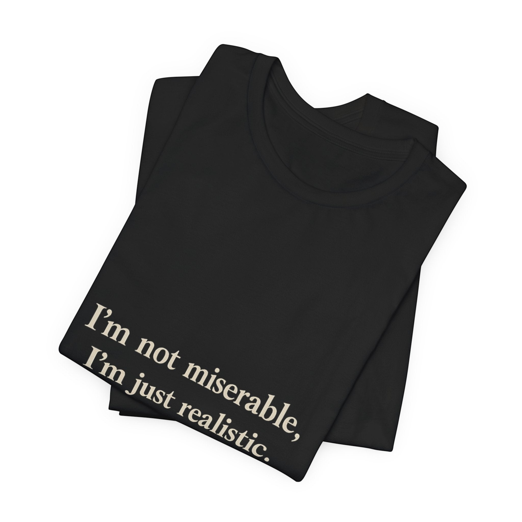 A folded black crew neck "I'm Not Miserable. I'm Just Realistic." T-shirt by Printify, branded Old Man Snark, with the slogan printed in light letters near the bottom corner.
