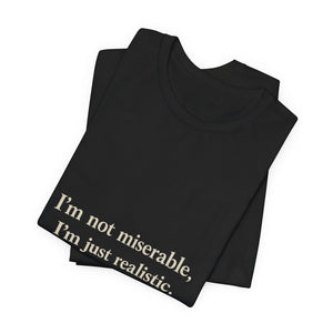 A folded black crew neck "I'm Not Miserable. I'm Just Realistic." T-shirt by Printify, branded Old Man Snark, with the slogan printed in light letters near the bottom corner.