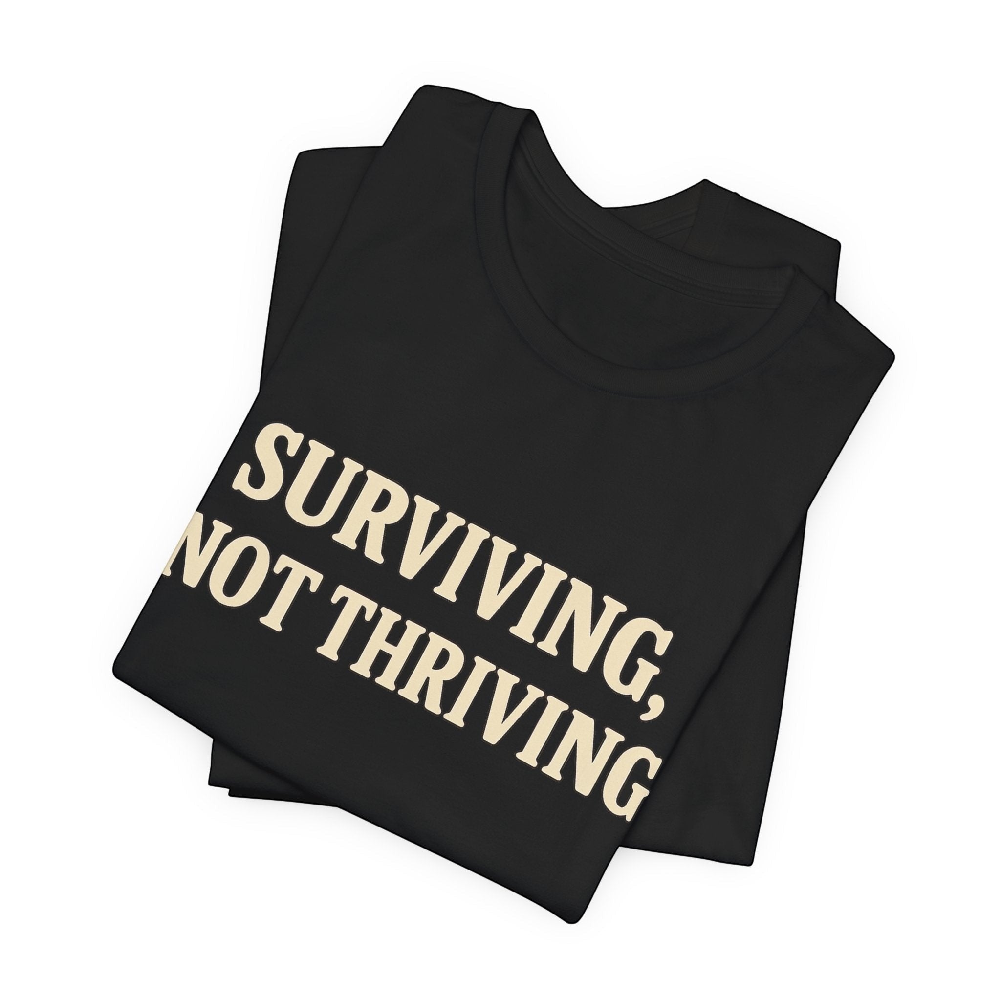 The "Surviving. Not Thriving." T-Shirt by Printify—Old Man Snark edition—is a folded black tee crafted from soft Airlume cotton, featuring bold beige lettering for an honest statement on modern life.