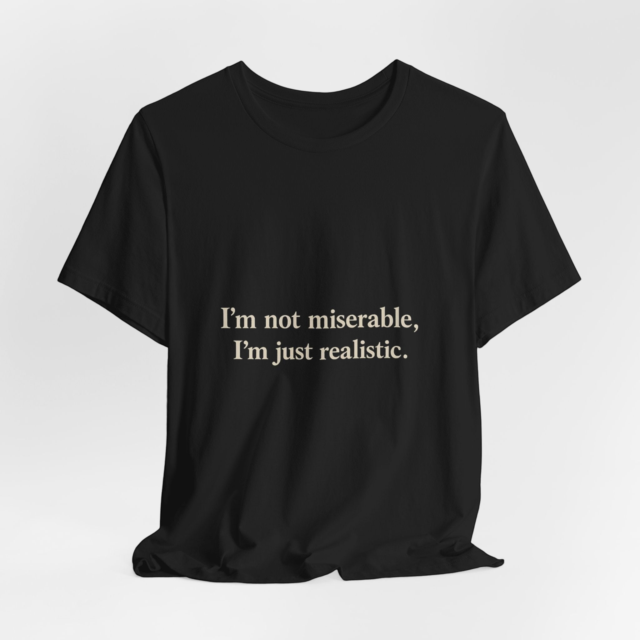 The "I'm Not Miserable. I'm Just Realistic." T-Shirt by Printify features bold white text on black Airlume cotton with a classic crew neck, shown against a plain white background.