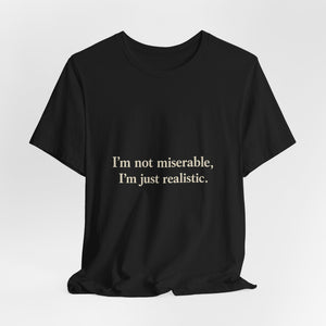 The "I'm Not Miserable. I'm Just Realistic." T-Shirt by Printify features bold white text on black Airlume cotton with a classic crew neck, shown against a plain white background.