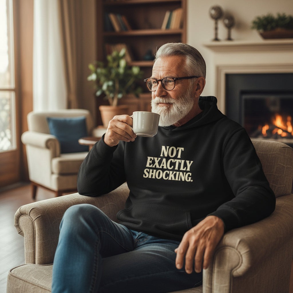 Old Man Snark black cotton-blend hoodie with 'NOT EXACTLY SHOCKING.' graphic text on chest, worn by a man sitting in an armchair.