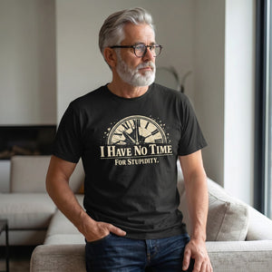 An older man with gray hair and a beard, wearing glasses and a Printify "I Have No Time For Stupidity" T-Shirt by Old Man Snark, sits on a couch in a modern living room.