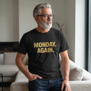 A gray-haired, bearded man in glasses wears the Printify "Monday. Again." T-Shirt by Old Man Snark as he sits thoughtfully on a modern beige couch near a window.