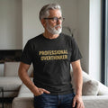 A gray-haired, bearded man in glasses sits on a bright couch, thoughtfully gazing out the window while wearing a Printify "Professional Overthinker" T-Shirt by Old Man Snark made from 100% Airlume cotton.