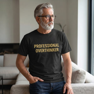 A gray-haired, bearded man in glasses sits on a bright couch, thoughtfully gazing out the window while wearing a Printify "Professional Overthinker" T-Shirt by Old Man Snark made from 100% Airlume cotton.