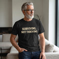 A gray-haired, bearded man in glasses wears the Printify "Surviving. Not Thriving." T-Shirt by Old Man Snark as he sits on a beige sofa in a sunlit living room, embodying modern life apparel.