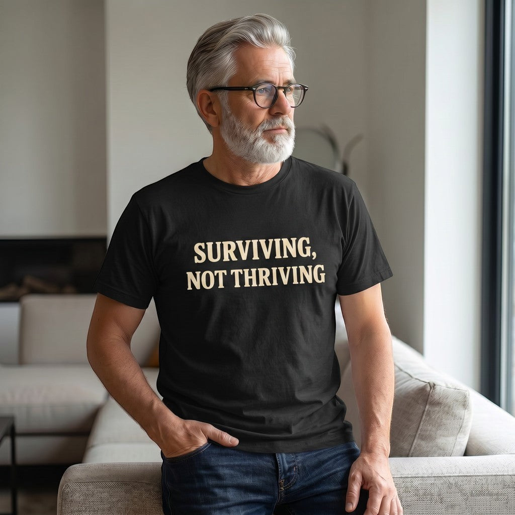A gray-haired, bearded man in glasses wears the Printify "Surviving. Not Thriving." T-Shirt by Old Man Snark as he sits on a beige sofa in a sunlit living room, embodying modern life apparel.