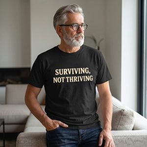 A gray-haired, bearded man in glasses wears the Printify "Surviving. Not Thriving." T-Shirt by Old Man Snark as he sits on a beige sofa in a sunlit living room, embodying modern life apparel.